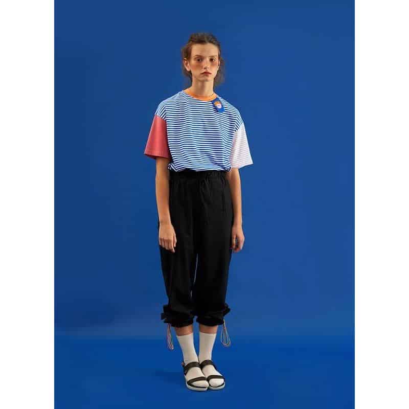 A young woman wearing a stylish, colorful striped T-shirt paired with black jogger pants, white socks, and trendy sandals, showcasing fashionable ophthalmic eyewear on a bright blue background.