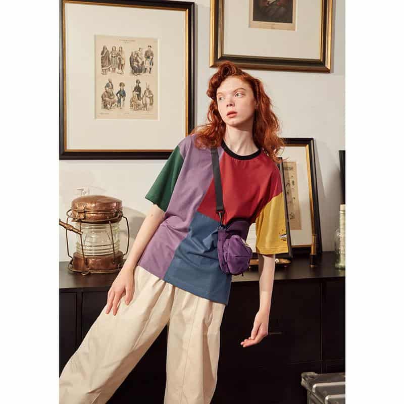 A young woman wearing a vibrant patchwork t-shirt and beige wide-leg pants accessorized with a purple waist bag, standing in a stylish interior setting with framed artwork. This trendy outfit combines casual comfort with colorful design, appealing to fashion-forward individuals seeking stylish everyday wear.
