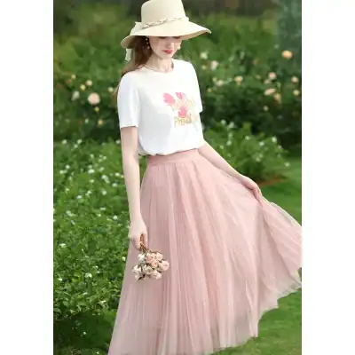 Beautiful woman wearing a pastel pink pleated skirt and white graphic T-shirt in a lush outdoor setting, showcasing fashionable women's clothing from Monalisa Store.