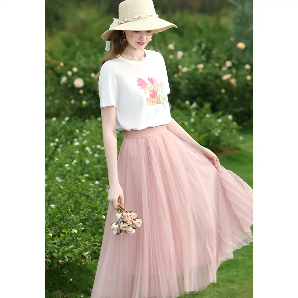 Beautiful woman wearing a pastel pink pleated skirt and white graphic T-shirt in a lush outdoor setting, showcasing fashionable women's clothing from Monalisa Store.
