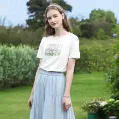 Stylish young woman wearing a comfortable white t-shirt with floral print and a flowy light blue skirt, accessorized with jewelry, standing outdoors in a lush garden setting for a Monalisa Store fashion photoshoot.