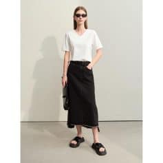 A stylish woman wearing a simple white V-neck T-shirt, black distressed denim maxi skirt, and chunky sandals, accessorized with black sunglasses and a clutch bag for a modern, casual look.