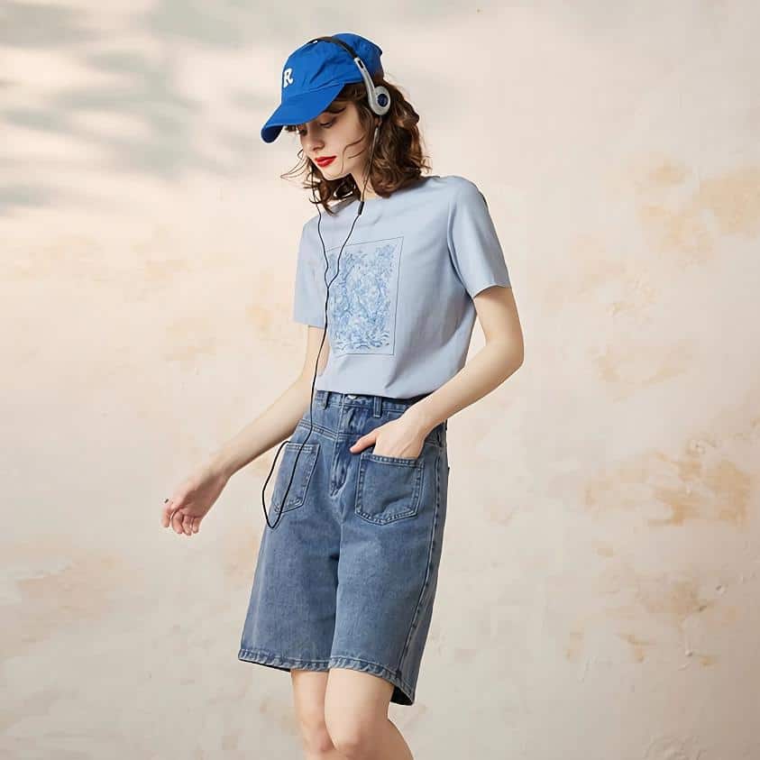 Woman listening to music with headphones, wearing casual summer outfit, denim shorts, and a cap, standing against a neutral background.