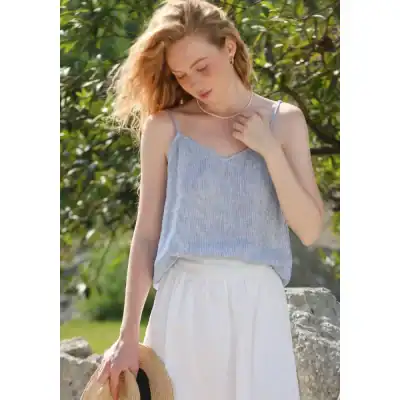 A young woman wearing a stylish, light blue summer top paired with a white skirt, holding a straw hat, enjoying sunny outdoor setting, perfect for summer fashion and stylish casual wear.