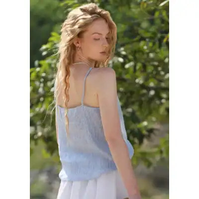 A woman in a light blue striped sleeveless top with thin straps, enjoying a sunny outdoor setting with lush green foliage in the background, perfect for casual summer fashion.