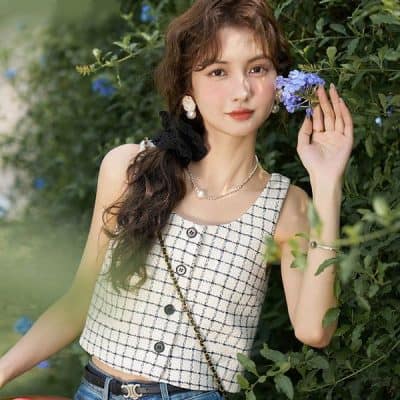 Beautiful young woman with wavy brown hair posing outdoors among lush greenery and vibrant blue flowers. Wearing a stylish checkered sleeveless top, jewelry, and accessories, embodying modern, chic fashion with a natural backdrop.