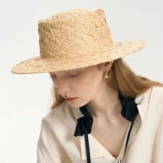 A woman wearing a natural straw hat, perfect for summer sun protection and outdoor fashion, complemented by elegant earrings and a beige blouse with dark accents.