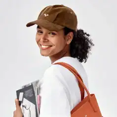 Close-up of a smiling woman with curly hair wearing trendy brown cap, white shirt, and orange tote bag, holding eyewear and sunglasses catalog, showcasing fashionable eyewear collection at Monalisa Store.
