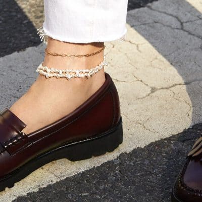 Close-up of a woman's ankle adorned with a pearl anklet, paired with a gold chain and white distressed jeans, showcasing fashionable jewelry suitable for summer and casual outings.