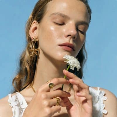 Close-up of a woman with clear skin, showcasing stylish eyeglasses and jewelry, holding a white flower against a blue background. Perfect setting for showcasing trendy optical accessories and eyewear styles.