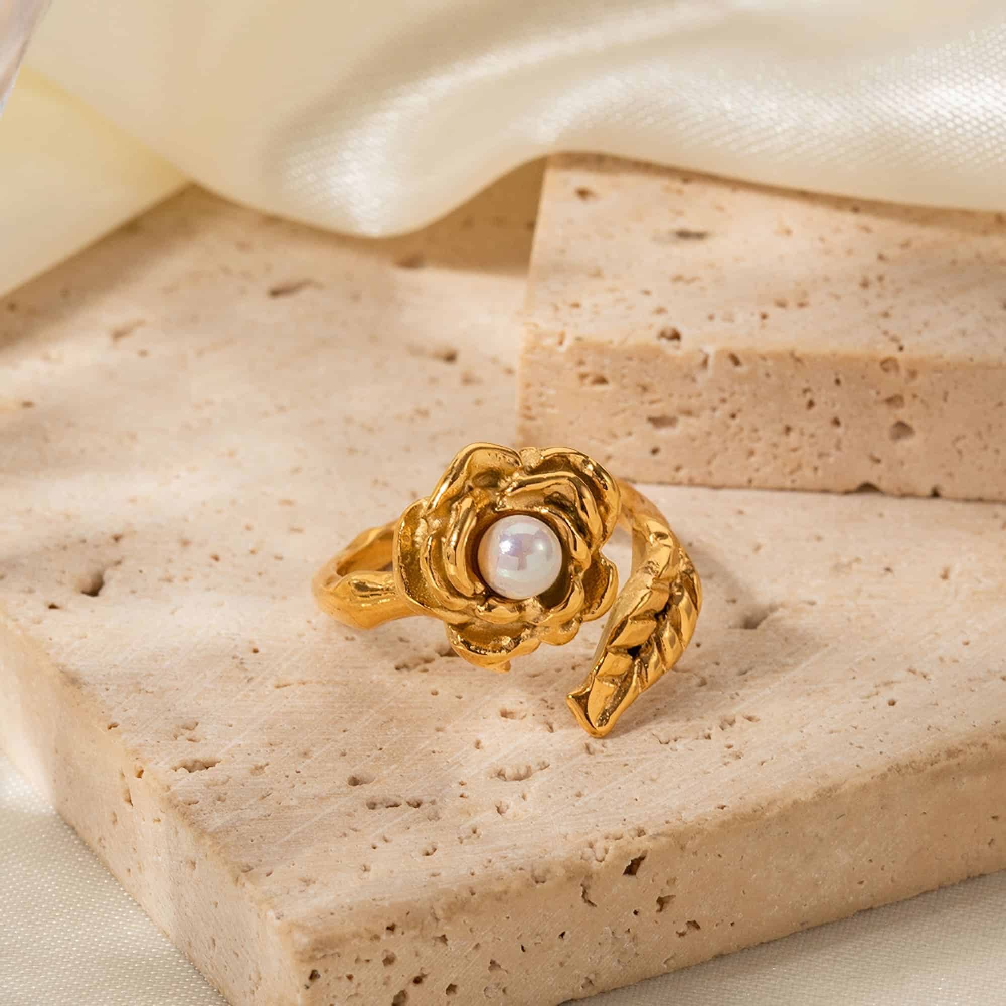 A stunning gold ring featuring a central pearl designed as a blooming flower, showcased on a textured beige surface with soft lighting. Perfect for luxury jewelry and sophisticated fashion accessories.