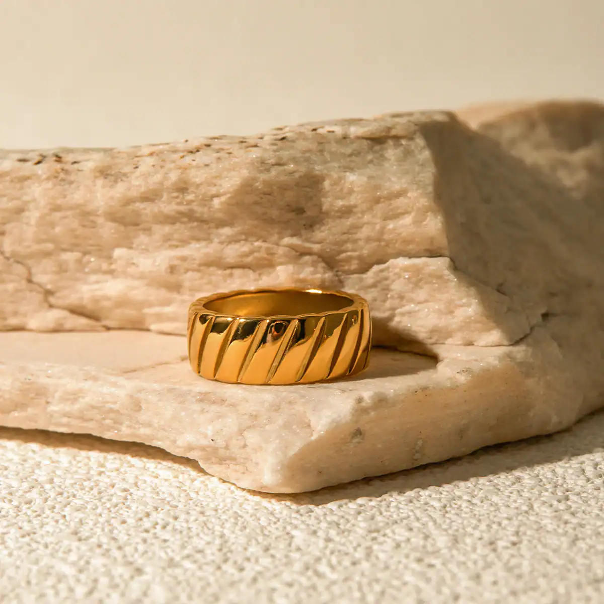 Close-up of a shiny gold ring with diagonal grooves, displayed on a textured stone surface for a luxurious jewelry presentation.