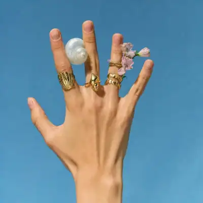 Close-up of a hand wearing multiple gold rings, adorned with pink flowers and a seashell, set against a clear blue sky, emphasizing jewelry and fashion accessories.