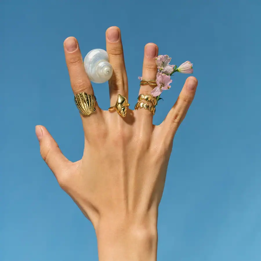 Close-up of a hand wearing multiple gold rings, adorned with pink flowers and a seashell, set against a clear blue sky, emphasizing jewelry and fashion accessories.