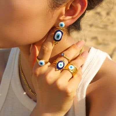 Close-up of stylish evil eye rings and earrings with blue and white designs, gold accents, on a woman's hand and ear, emphasizing jewelry fashion and accessories at Monalisa Store.