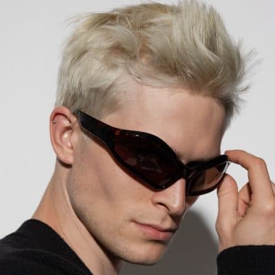 Close-up of a young man with platinum blonde hair, wearing dark sunglasses, showcasing modern fashion accessories and a youthful, stylish look.