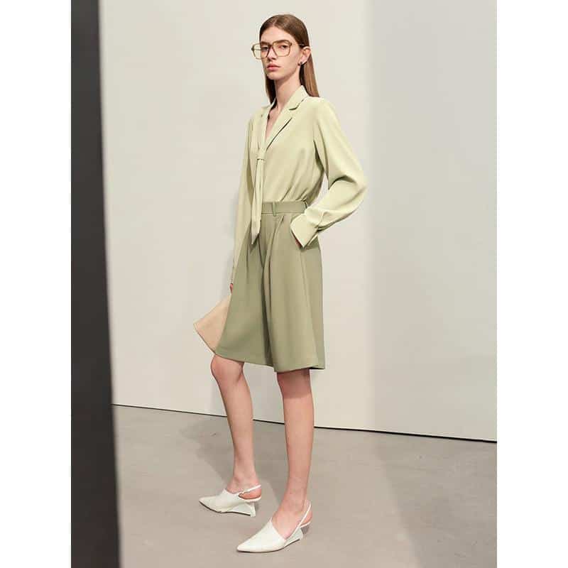 Stylish woman wearing a professional light green blazer and matching knee-length shorts, paired with white heels and glasses, showcasing modern office fashion.