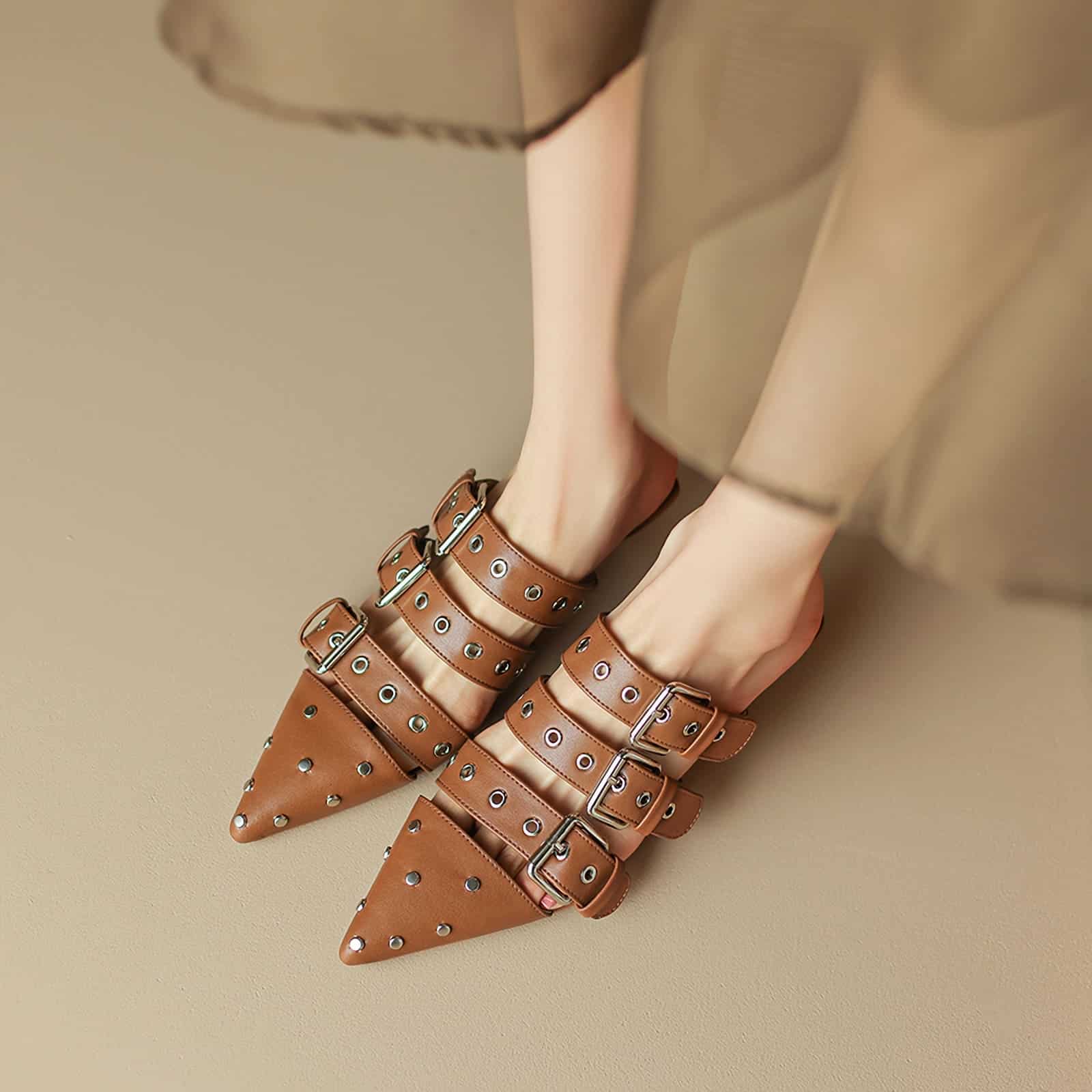 Close-up of stylish brown pointed-toe shoes with multiple buckles and metallic eyelets, designed for a chic, trendy look.