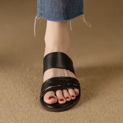 Close-up of stylish black women's sandals with a thick strap and open toes, featuring red nail polish on toes, worn with ripped jeans, on beige carpet floor, showcasing trendy footwear options from Monalisa Store.