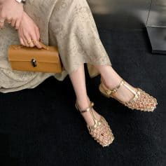 Fashionable gold sandals with intricate netted straps and ankle buckles, perfect for chic occasions, paired with a beige floral dress and matching accessories.