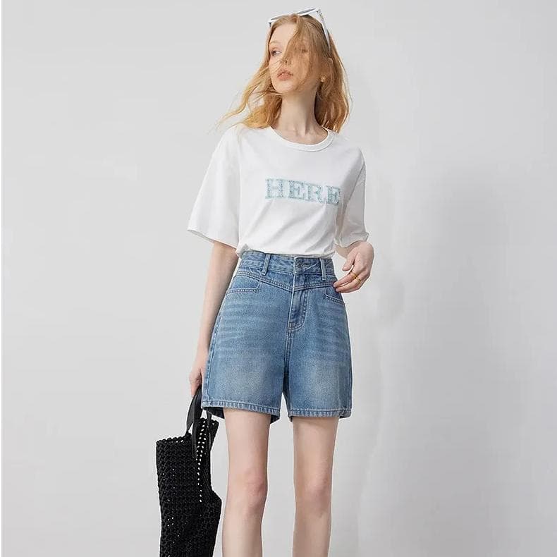 A young woman wearing a white T-shirt with "HERE" written in blue, paired with high-waisted denim shorts. She holds a black woven tote bag, highlighting trendy and comfortable fashion essentials.