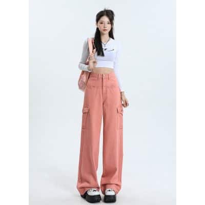 A young woman with long dark hair dressed in trendy oversized pink cargo pants, white crop top, and chunky shoes, posing confidently against a plain white background.