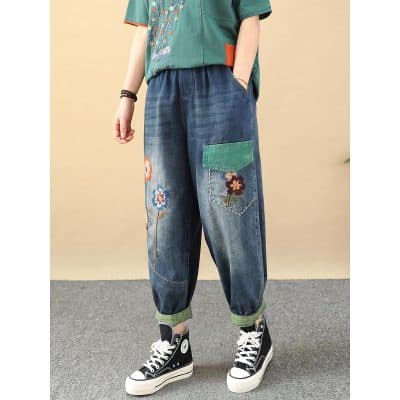 A pair of trendy denim pants featuring vibrant floral embroidery, a green patch pocket, and a relaxed fit with rolled-up cuffs, perfect for a casual and stylish look.