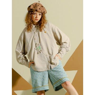 A young woman modeling trendy casual wear, including a beige fluffy jacket, blue gingham shorts, and a brown beret with polka dots, perfect for fashion-forward, comfortable everyday style.