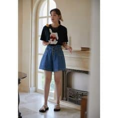 A young woman wearing a casual black t-shirt with a bunny graphic, high-waisted denim shorts, and sandals, standing inside a bright, elegant room with a fireplace and arched window.