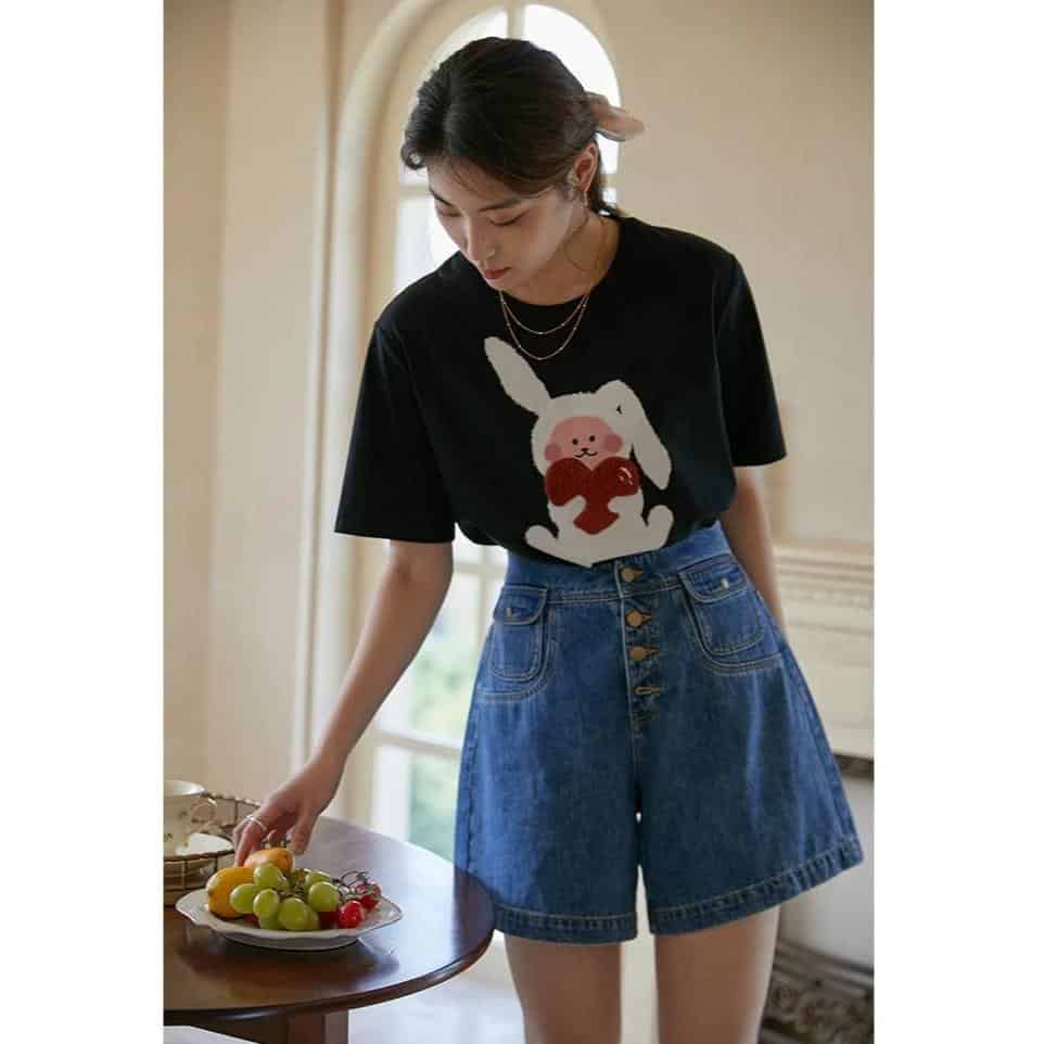 A young woman wearing a black casual T-shirt with a cute bunny graphic holding a heart, styled with blue denim shorts, showcasing playful and fashionable attire from Monalisa Store. Perfect for casual wear and everyday fashion.