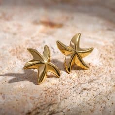 Shiny gold starfish earrings resting on textured sand, perfect for ocean-themed jewelry lovers and beach fashion accessories.