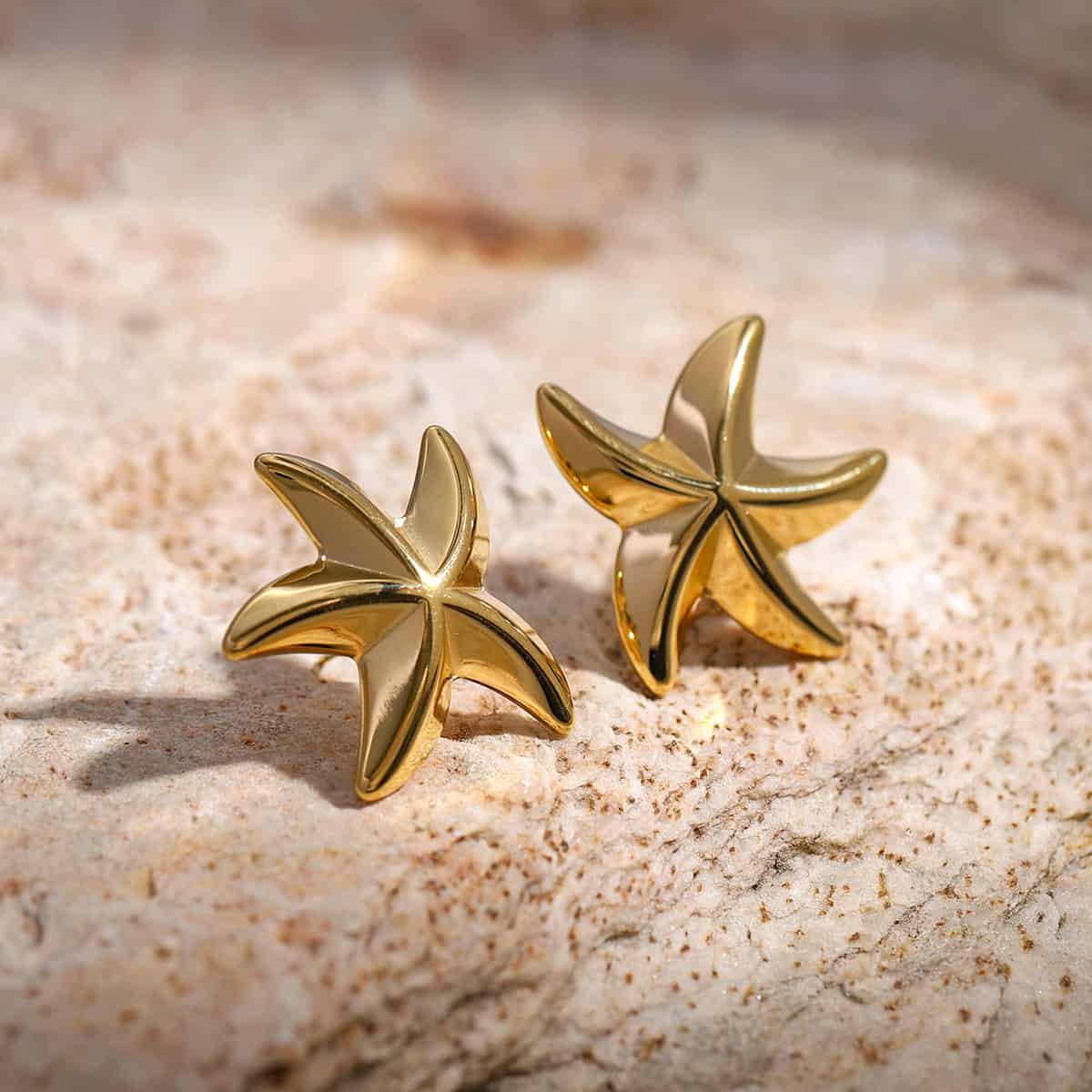 Shiny gold starfish earrings resting on textured sand, perfect for ocean-themed jewelry lovers and beach fashion accessories.