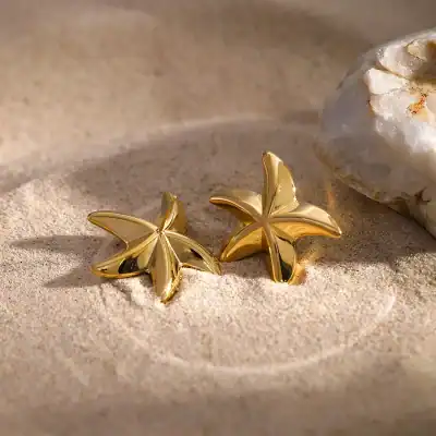 Delicate gold butterfly earrings resting on sand, perfect for summer accessories and beachwear. Ideal for stylish women seeking unique jewelry for casual or special occasions.