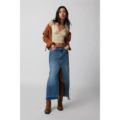 Stylish young woman wearing a long denim skirt with a front slit, paired with a beige lace top, brown leather boots, and a rust-colored leather jacket, showcasing trendy casual fashion.