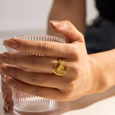 Close-up of a gold ring featuring detailed craftsmanship, worn on a finger holding a textured glass cup, showcasing luxury jewelry styles.