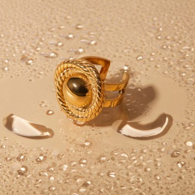 Elegant gold ring with a black pearl centerpiece, featuring intricate twisted detailing, showcased on a sandy beach with shells and bubbles.