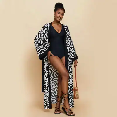 Stylish model wearing a black swimsuit with a fashionable zebra-print cover-up, accessorized with black strappy heels and a woven handbag for a chic summer look.