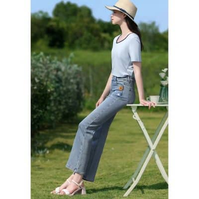 Stylish woman sitting on garden table with greenery background, wearing light blue jeans, summer top, straw hat, and sandals, emphasizing fashion, comfort, and outdoor leisure.