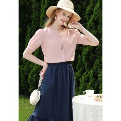Bright image of a young woman outdoors wearing a pink blouse, navy skirt, straw hat, and jewelry, highlighting stylish summer fashion and accessories.