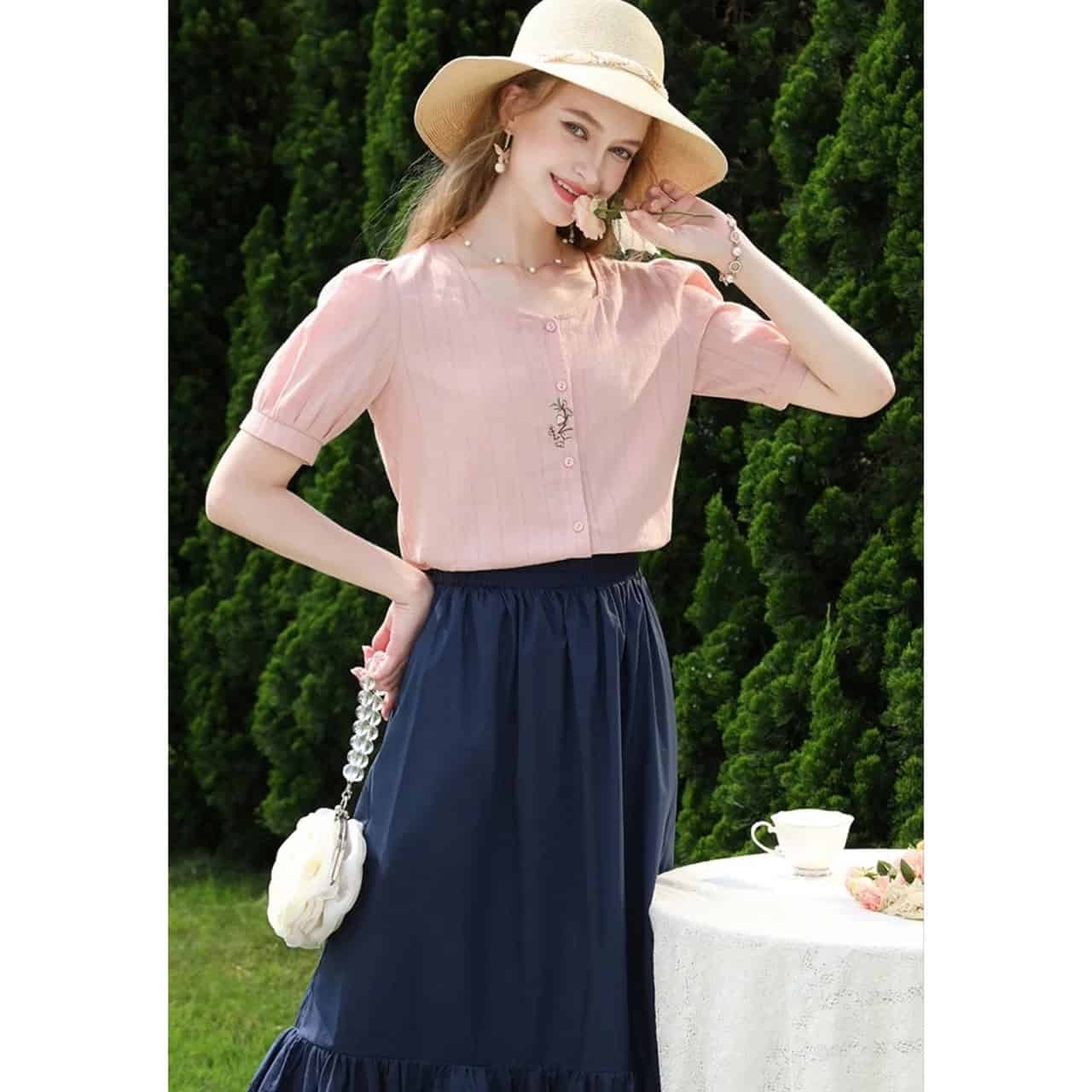 Bright image of a young woman outdoors wearing a pink blouse, navy skirt, straw hat, and jewelry, highlighting stylish summer fashion and accessories.