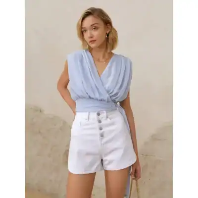Fashionable light blue sleeveless wrap top paired with high-waisted white denim shorts featuring button-front closure, perfect for trendy summer outfits and casual wear.