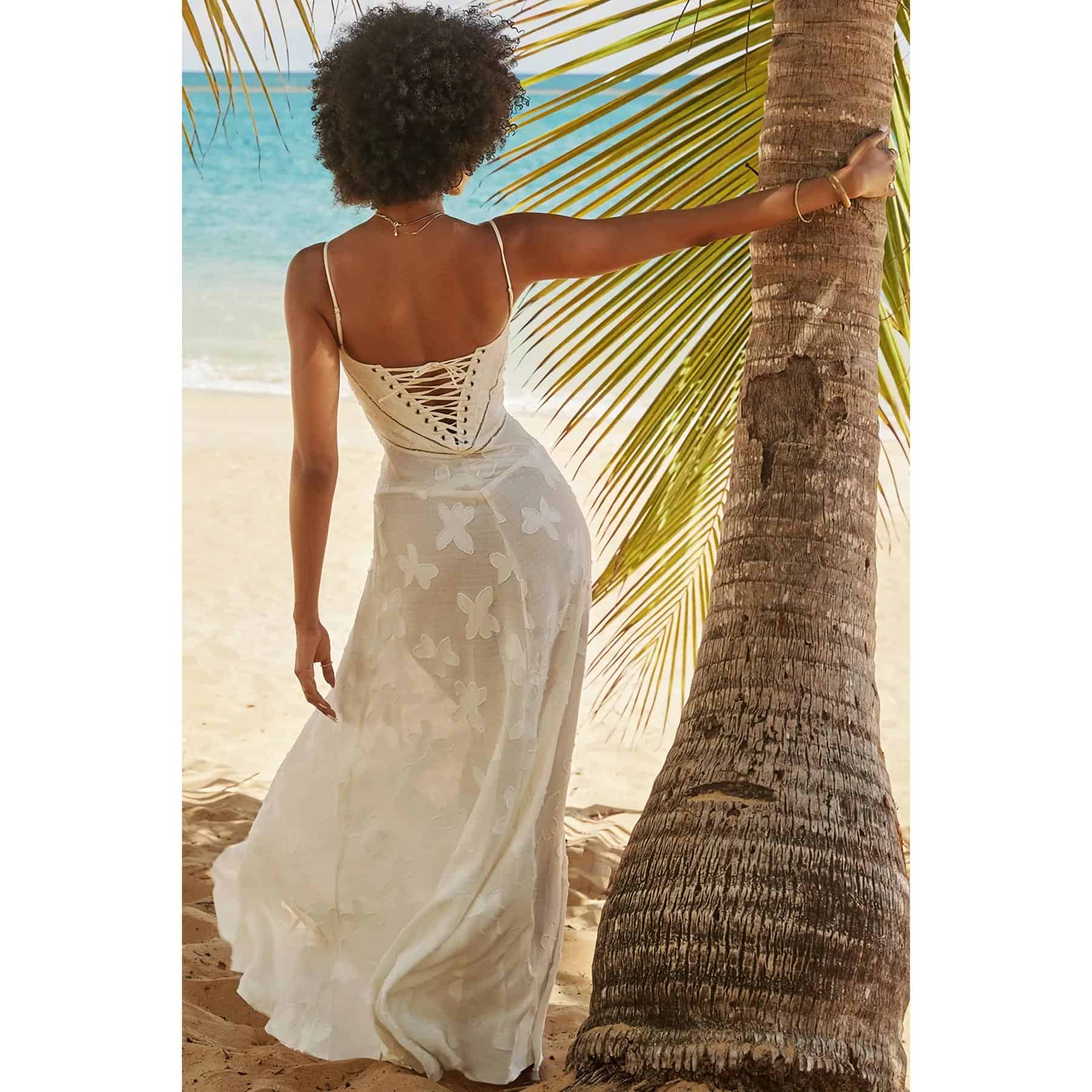 A woman in a white, butterfly-embroidered dress on the beach, standing next to a palm tree, showcasing stylish resort fashion and summer clothing available at Monalisa Store.