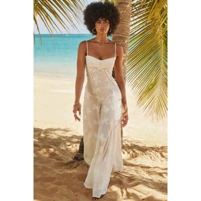 Beautiful woman in a stylish white floral dress on a tropical beach with palm trees and ocean backdrop.