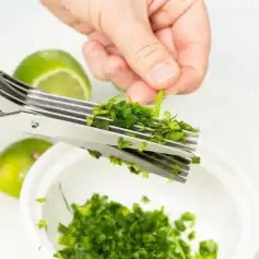 Close-up of a hand finely chopping green chives with a knife over a white bowl, with lime slices in the background, emphasizing fresh ingredients for healthy meals.
