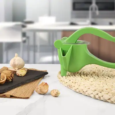 A bright green garlic press placed on a woven mat with garlic cloves and slices nearby, ideal for kitchen use and food preparation at Monalisa Store.