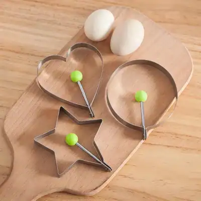 Cookie cutters in heart and star shapes with green handles, suitable for baking and kitchen use, placed on a wooden board with eggs in the background.