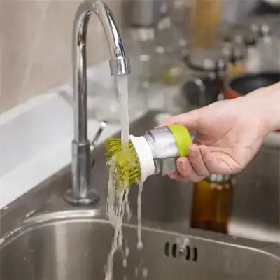 A person rinsing a green and white facial cleansing brush under running water in a kitchen sink for proper hygiene.