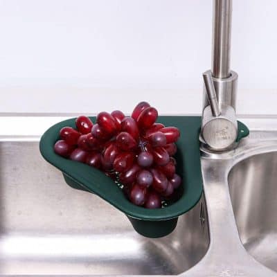 Fresh red grapes placed in a green colander on a stainless steel kitchen sink with modern faucet.