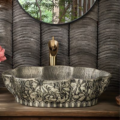 A luxurious marble bathroom sink with floral carved details, paired with a modern gold faucet, set against textured dark wall panels and a large round mirror reflecting lush greenery.
