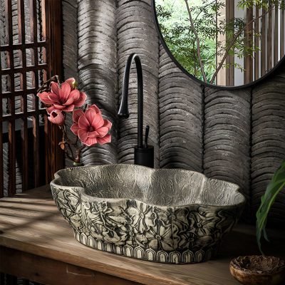A decorative bathroom sink with carved stone bowl, black faucet, floral arrangement, and natural elements for a luxury spa feel.